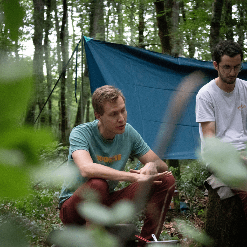 Rémi Lede assis autour d'un feu de camp en forêt, accompagné de plusieurs participants, entourés d'arbres et de la nature.